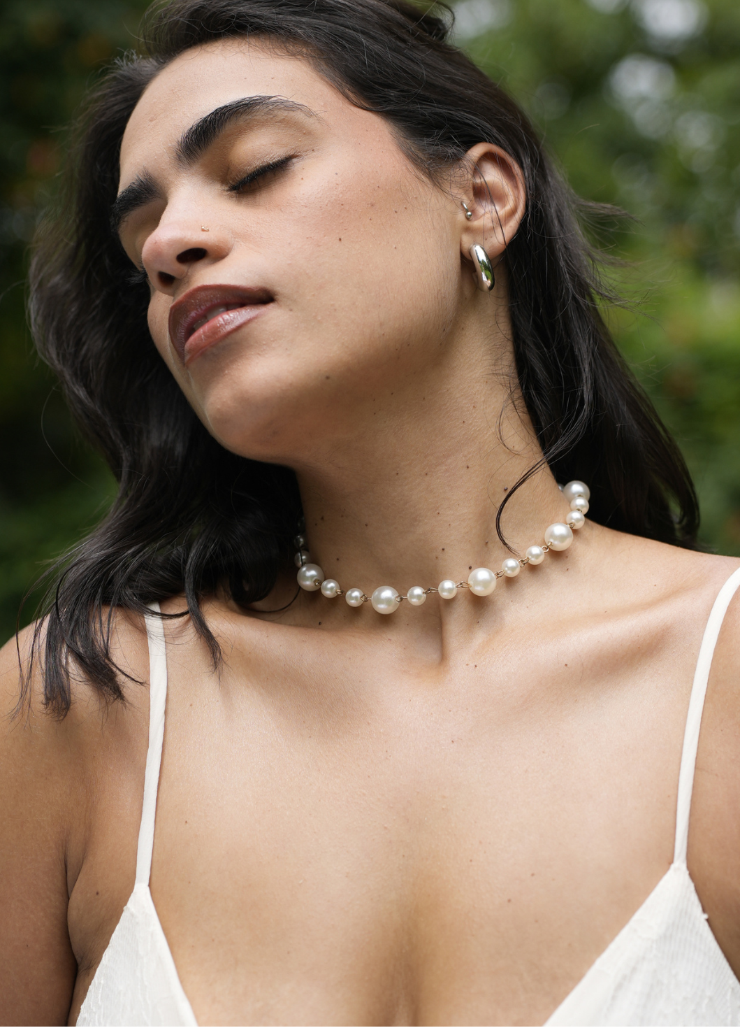 A woman outdoors wearing a pearl choker necklace and a matching pearl stud earring, with green foliage blurred in the background.